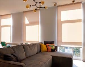 Motorized roller blinds. A sofa with colorful pillows in the room near windows with roller blinds. Automatic roller shades on large window to the floor in the interior. Sunny day.
